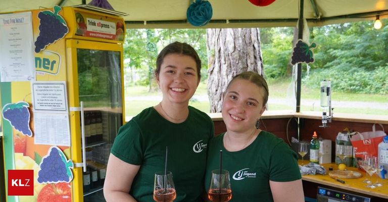 Zwei Frauen in grünen T-Shirts halten Weingläser. Sie stehen neben einem Getränkeautomaten mit einem Baum und dekorativen Gegenständen im Hintergrund.