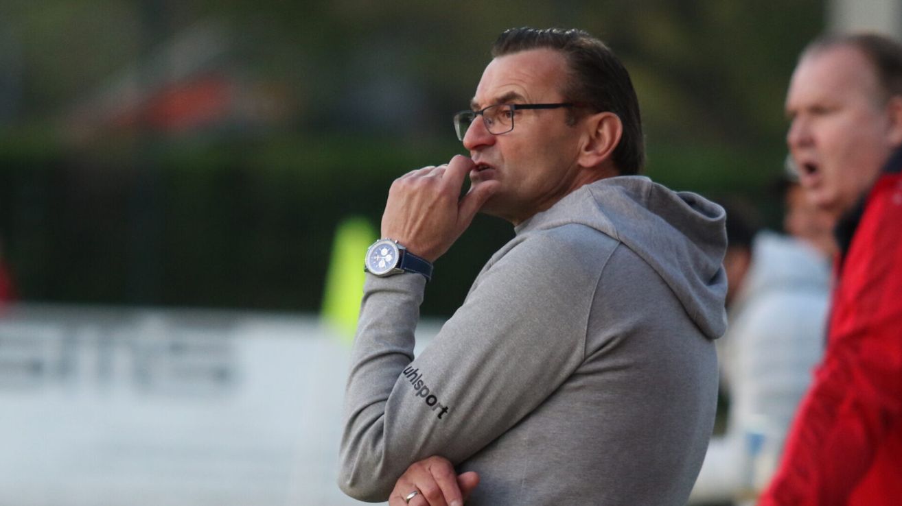 A man wearing a gray hoodie and glasses is standing outdoors, his hand on his chin. He wears a watch with a blue strap.