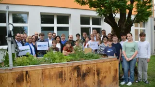 Eine Gruppe von Menschen posiert für ein Foto vor einem Gebäude mit einem hölzernen Pflanzgefäß und einem Baum. Einige halten Schilder.