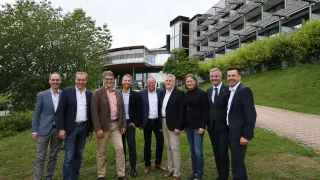 A group of formally dressed individuals stand on a lawn, smiling for a photo in front of a modern building with glass walls.