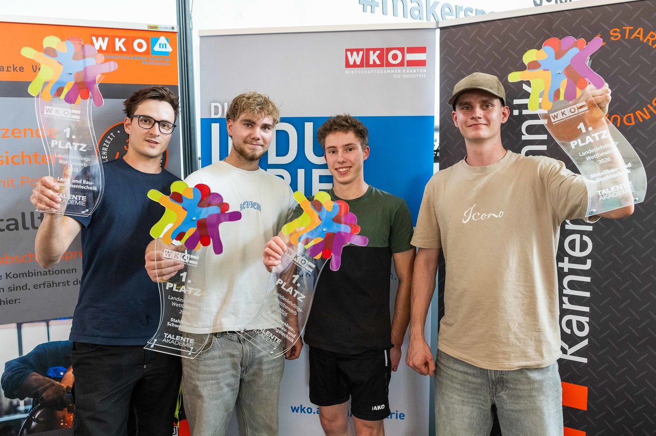 Vier junge Männer stehen lächelnd zusammen und halten Trophäen, wahrscheinlich bei einem Wettbewerb. Hinter ihnen ist ein Banner mit 'WKO' und 'DU BRIE'. Eine Trophäe steht '1. Platz'.