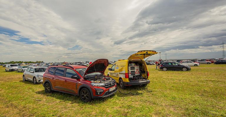 Ein roter SUV mit offenem Motorraum und ein gelber Servicewagen mit offener Heckklappe sind auf einem Rasenfeld geparkt.