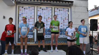 Four athletes stand on a podium, smiling, with trophies in hand. They are wearing sportswear and have bib numbers on their shirts. Behind them, a banner displays various logos and dates.