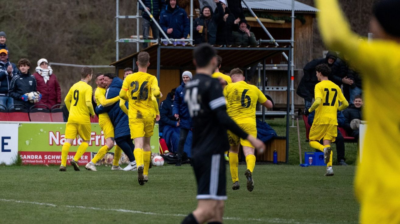 Soccer players in yellow uniforms celebrate on the field. Spectators in the stands watch, some cheering. A player in black stands in the foreground.