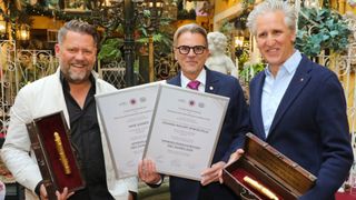 Three men in formal attire pose with certificates and a trophy. The man on the left holds a certificate labeled 'Spargelfeinschmecker des Jahres 2026'. The man in the middle holds a certificate with a gold-plated pen. The man on the right smiles. Behind them, a statue and decorative plants are visible.