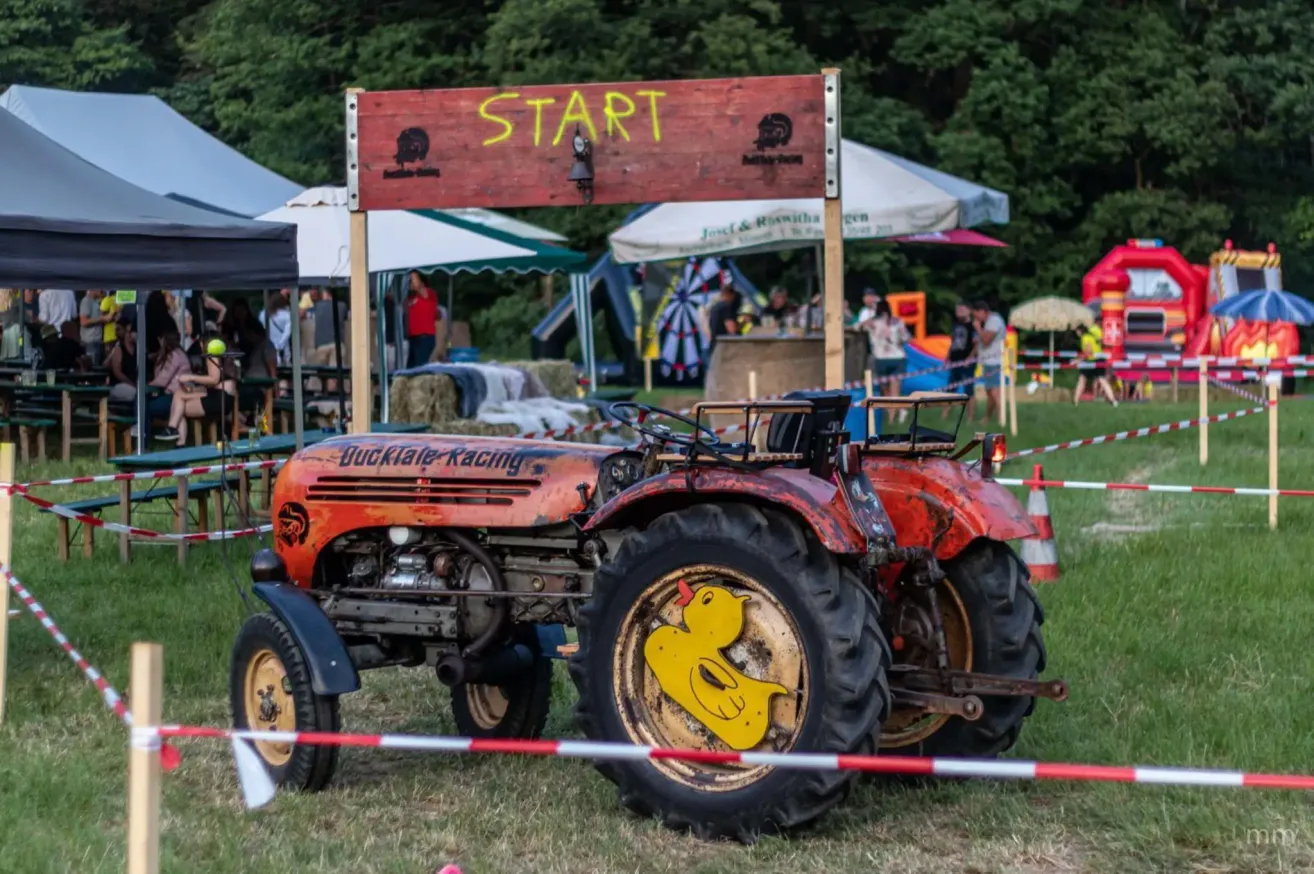 Ein alter roter Traktor mit einem Schild 'Start' und einer gelben Gummiente auf dem Reifen steht auf einem Feld mit Menschen und Zelten im Hintergrund.