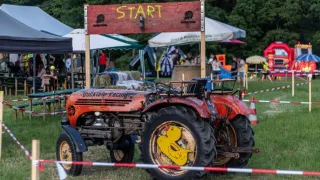 An old red tractor with a sign reading 'Start' and a yellow rubber duck on the tire is in a field with people and tents in the background.