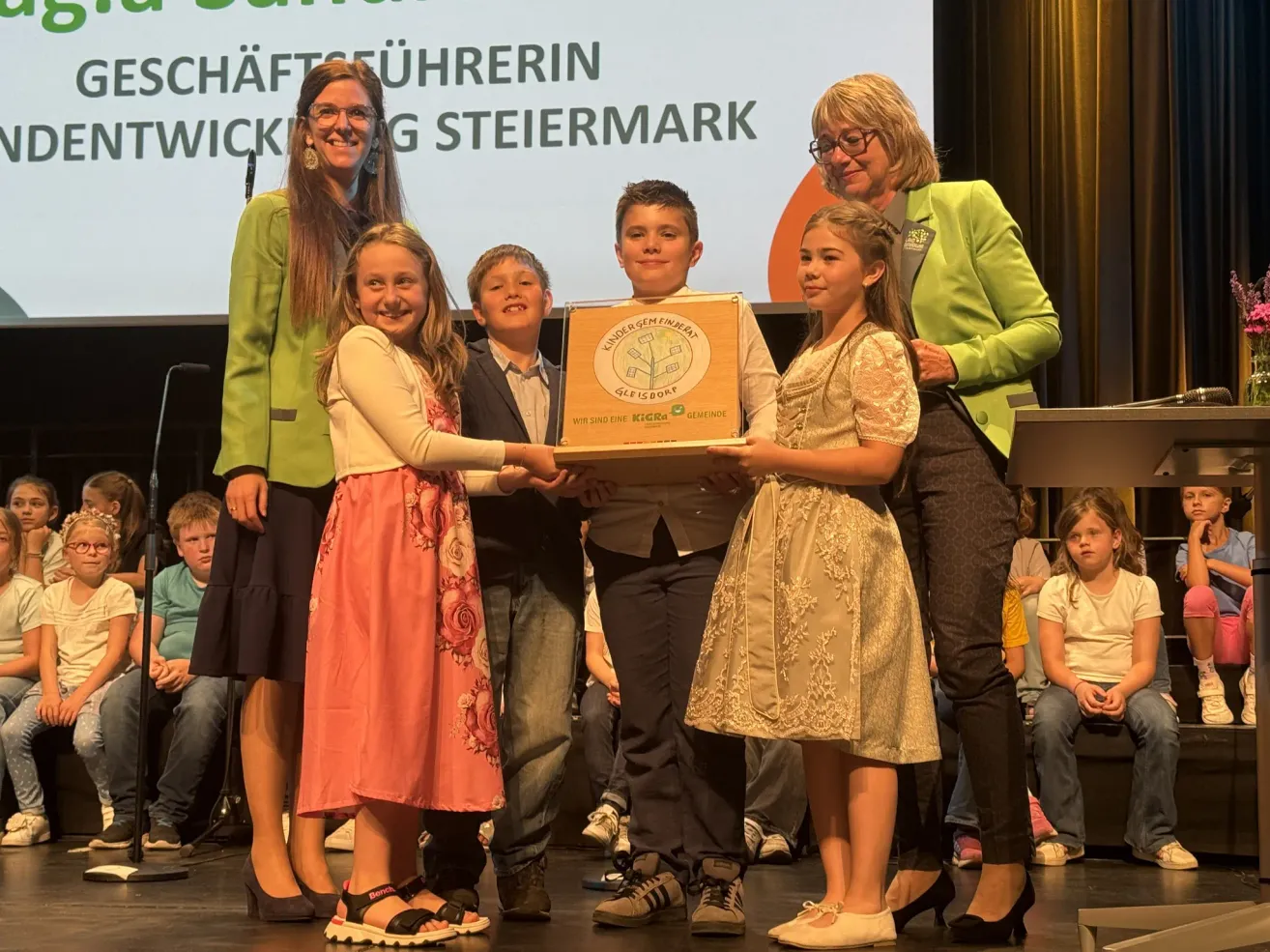 Mehrere Kinder und Erwachsene stehen auf der Bühne und halten eine Schachtel mit der Aufschrift "Kindergarten Steiermark". Sie lächeln und schauen nach vorne. Hinter ihnen beobachten zwei Frauen und ein Mann lächelnd. Die Kinder tragen Kleider und Turnschuhe, während die Erwachsenen Brillen und Lederschuhe tragen.
