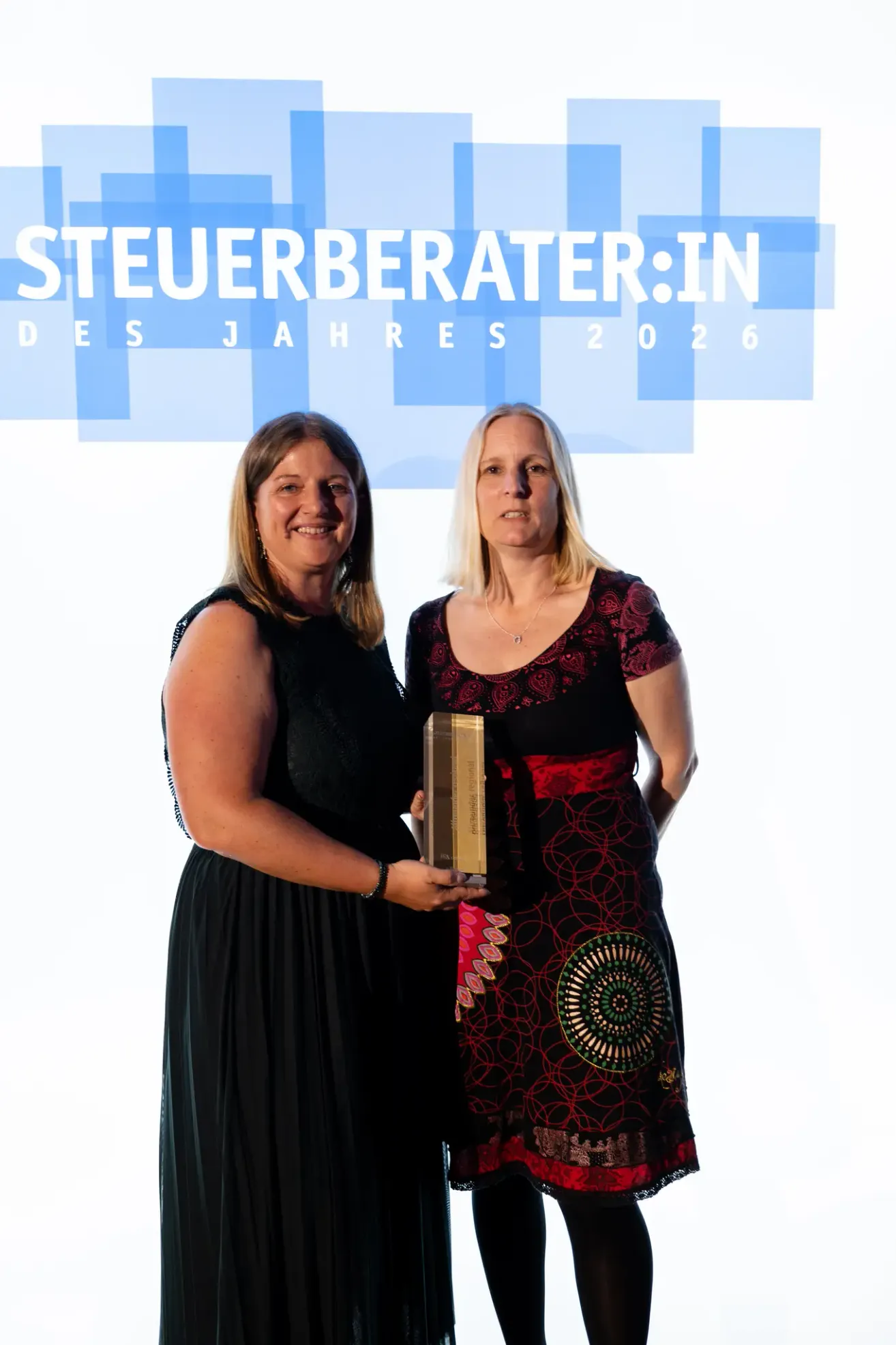 Zwei Frauen stehen vor einem blauen Banner mit der Aufschrift Steuerberater:in des Jahres 2026 und lächeln, während sie einen Preis halten.