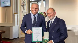 Two men in suits and ties stand in a room with a flag and awards. One man holds a certificate and a medal, while the other smiles. A flag, window, and a vase with flowers are in the background.