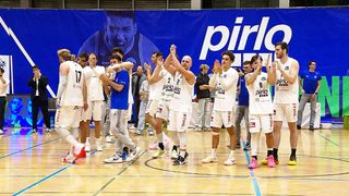 Basketballspieler feiern auf dem Spielfeld und stehen in weißen Trikots mit Nummern und Sponsoren. Im Hintergrund befindet sich ein blauer Banner mit dem Wort pirlo.