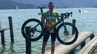 A man with a medal stands on a dock by the water, holding a bicycle. In the background, mountains and sailboats are visible.