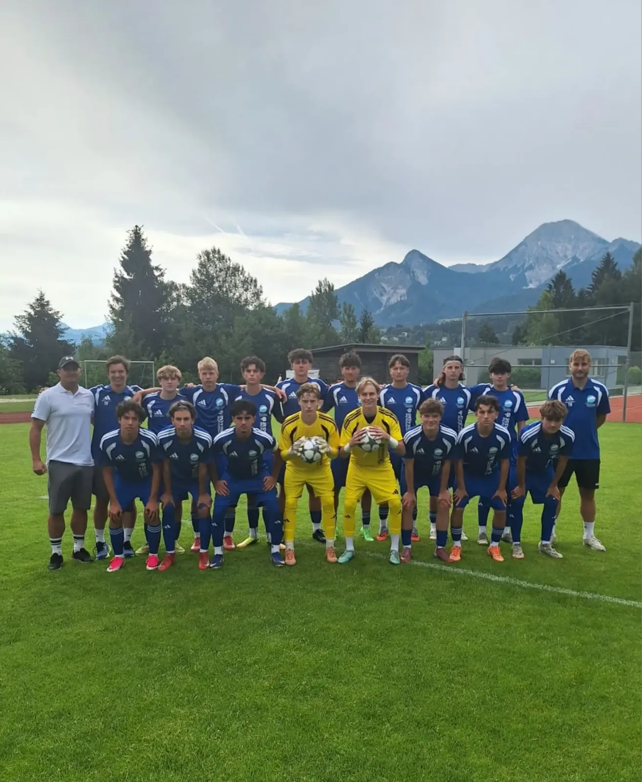 A soccer team in blue uniforms poses for a photo on a field with mountains in the background.