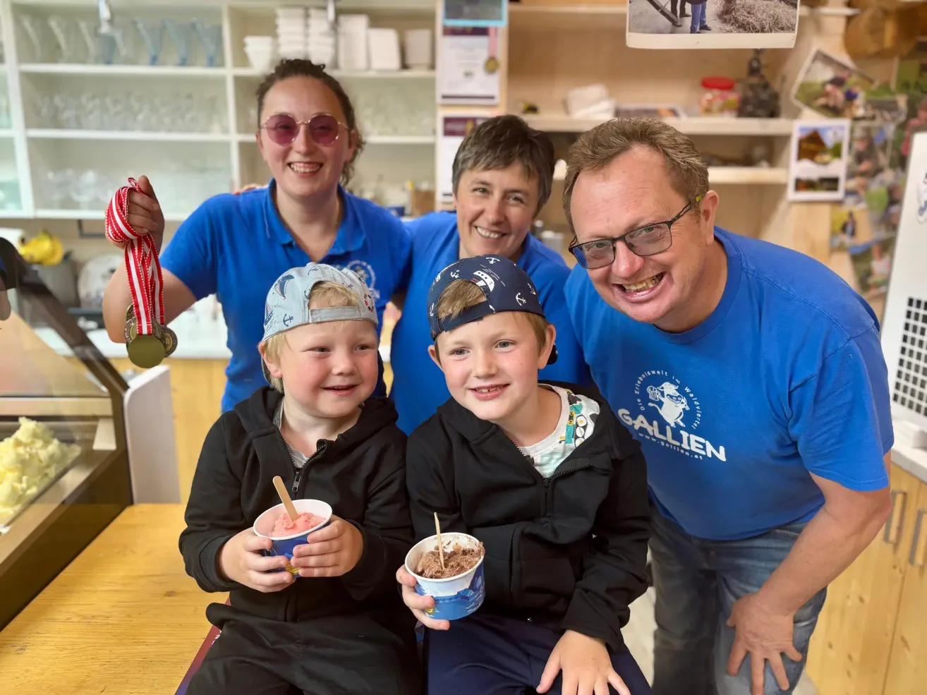 Vier Personen posieren für ein Foto. Zwei halten Eisbecher in der Hand. Die Frau hält eine Medaille. Der Mann hat ein lächelndes Gesicht. Sie tragen alle blaue Hemden.
