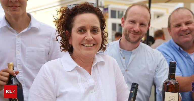 A woman with curly hair smiles while holding a wine bottle. A man stands beside her, smiling, in an outdoor setting.
