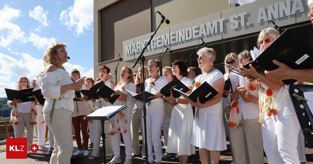 Ein Chor von Frauen in weißen Kleidern und Schals steht vor einem Gebäude, singt und hält Musikbücher und Mikrofone in den Händen.