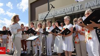 Ein Chor von Frauen in weißen Kleidern und Schals steht vor einem Gebäude, singt und hält Musikbücher und Mikrofone in den Händen.
