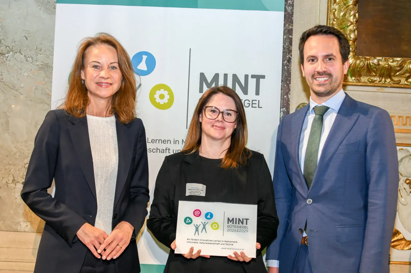 Drei Personen stehen vor einem Banner. Die Frau in der Mitte hält ein Zertifikat. Das Banner zeigt 'MINT' und Symbole der Wissenschaft. Die Personen sind formell gekleidet.