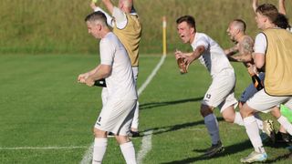 Zwei Männer feiern auf einem Fußballfeld, einer öffnet eine Flasche. Ein dritter Mann hebt einen Ball hoch, während ein weiterer hinter ihnen steht.