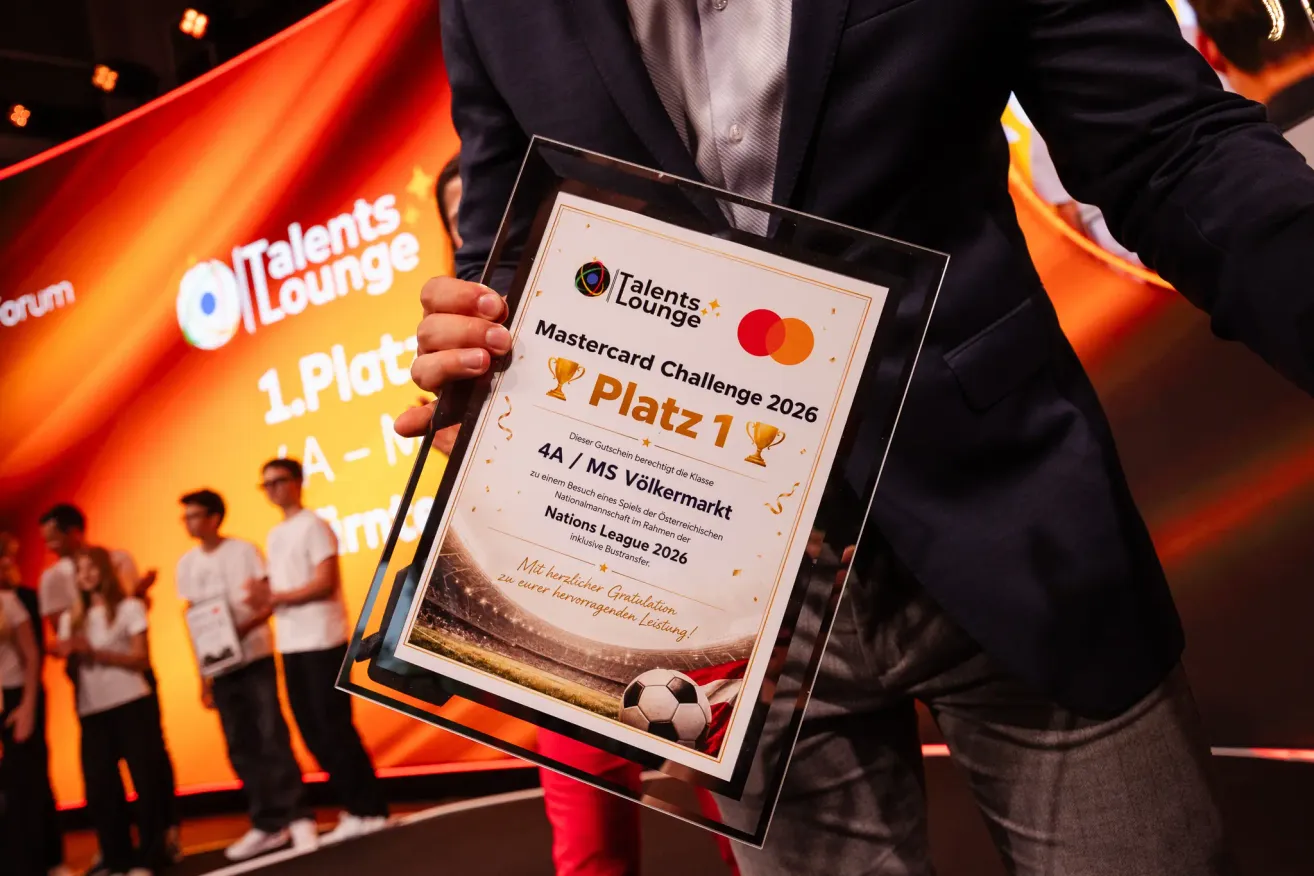 Ein Mann hält eine Glasplakette mit einem Fußball darauf. Es steht Platz 1 Mastercard Challenge 2026. Der Hintergrund ist ein Banner mit orangen und weißen Farben.