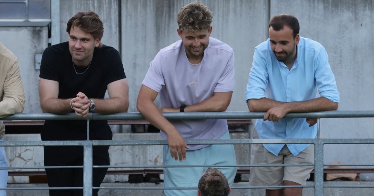 Drei Männer stehen auf einer Balkonbrüstung und lächeln. Der Mann in der Mitte trägt ein weißes T-Shirt und blaue Shorts. Der Mann auf der linken Seite trägt ein schwarzes T-Shirt und hat eine Uhr und eine Halskette. Der Mann auf der rechten Seite trägt ein blaues Hemd und hat eine Uhr.