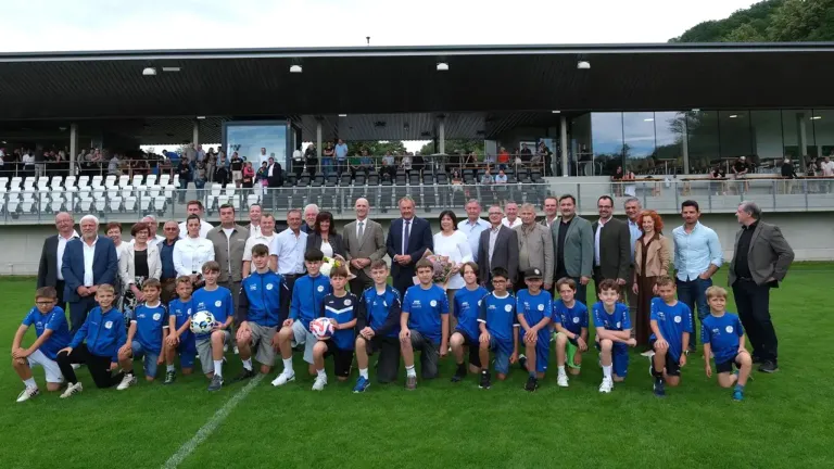 Eine Fußballmannschaft in blauen Uniformen posiert für ein Foto auf einem Spielfeld, mit Erwachsenen in Anzügen und Krawatten hinter ihnen. Einige halten Blumen und stehen auf den Tribünen.