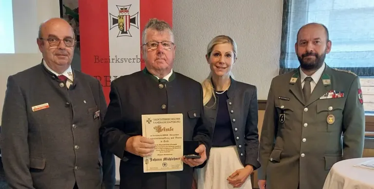 Eine Preisverleihung, bei der ein Mann und eine Frau eng beieinanderstehen. Der Mann hält ein Zertifikat und eine kleine Schachtel, während die Frau lächelt. Dahinter ist eine Kulisse mit einem Logo.