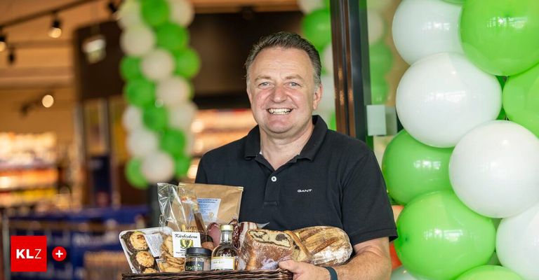 Ein Mann in einem schwarzen Poloshirt lächelt, während er einen Korb mit verschiedenen Lebensmitteln und einer Flasche hält, vor grünen Luftballons stehend.