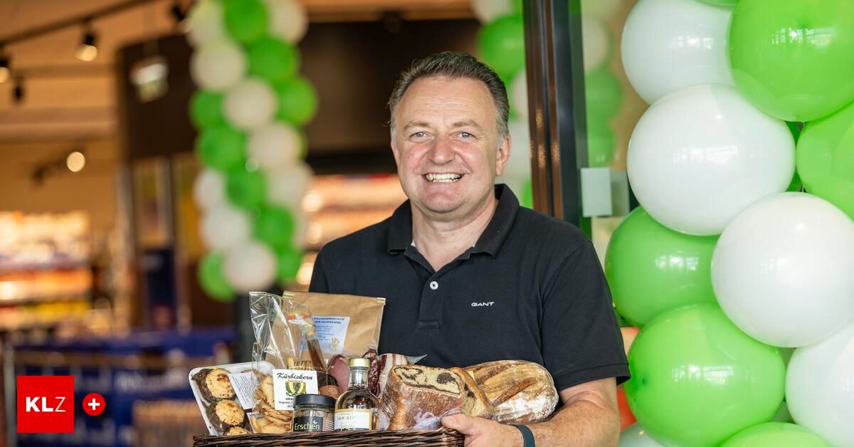 Ein Mann in einem schwarzen Poloshirt lächelt, während er einen Korb mit verschiedenen Lebensmitteln und einer Flasche hält, vor grünen Luftballons stehend.