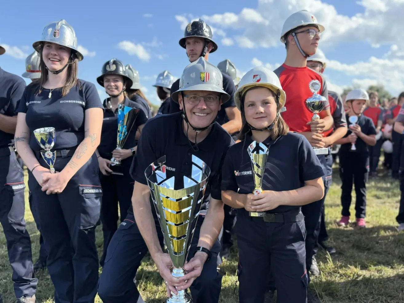 Eine Gruppe von Feuerwehrleuten in Uniform, die Pokale halten, lächeln und für ein Foto posieren. Ein Mann und ein Kind sind in der Mitte und halten beide große goldene Pokale.