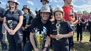 Eine Gruppe von Feuerwehrleuten in Uniform, die Pokale halten, lächeln und für ein Foto posieren. Ein Mann und ein Kind sind in der Mitte und halten beide große goldene Pokale.