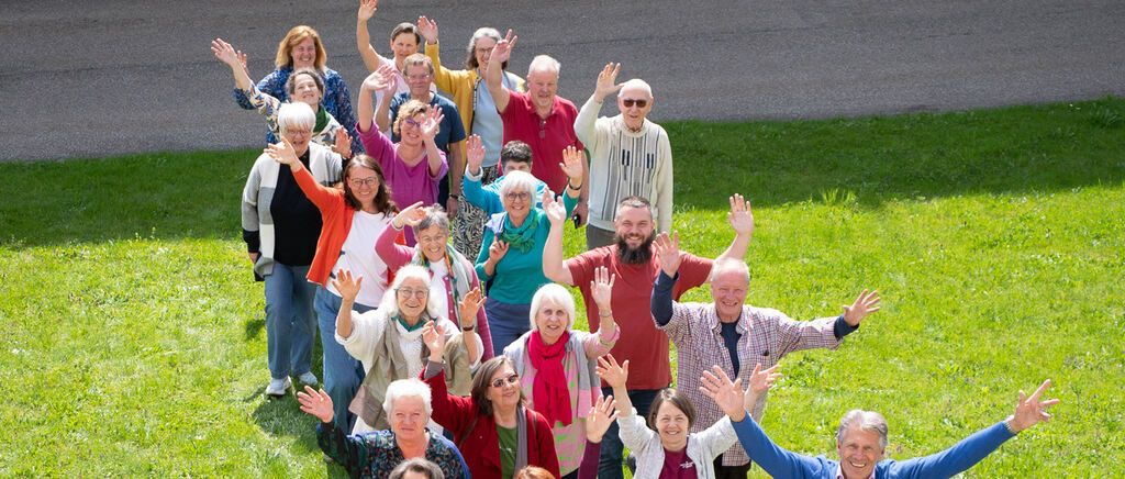A group of adults and seniors smiling and waving in a grassy field with a road in the background.