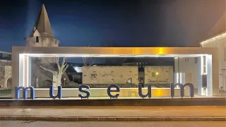 Ein Stadtmuseum ist nachts beleuchtet, in der Nähe steht ein Baum und eine Straßenlampe. Hinter dem Museum befindet sich ein Gebäude mit einer Wandmalerei.