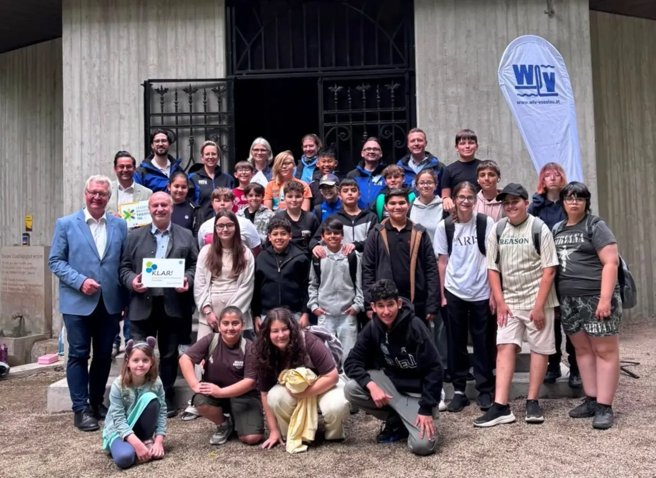 Eine Gruppe von Menschen, einschließlich Erwachsener und Kindern, posiert für ein Foto vor einem Gebäude mit einem Banner. Einige tragen Brillen, Mützen und Sneakers, während zwei ein Schild halten.