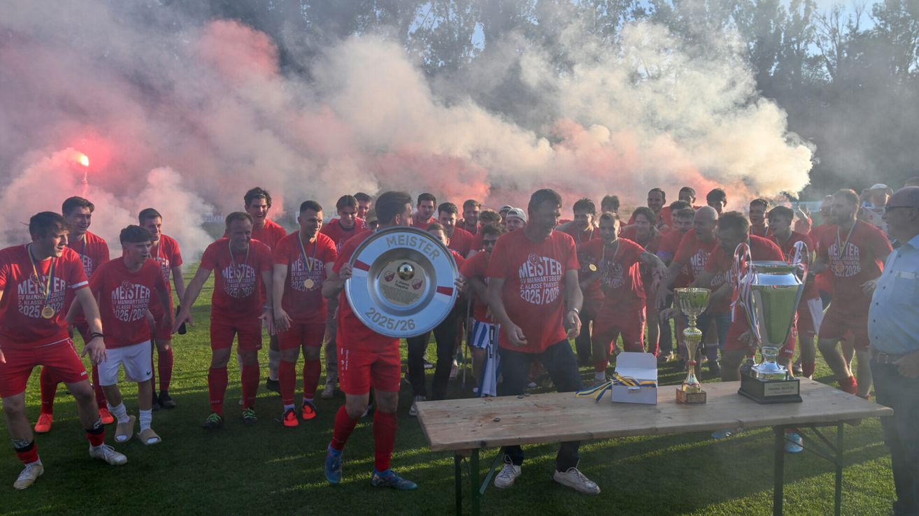 Eine Sportmannschaft feiert mit Trophäen auf einem Feld, umgeben von rotem und weißem Rauch. Ein Mann in der Mitte hält eine große silberne Trophäe, während ein anderer Mann eine goldene Trophäe hält.