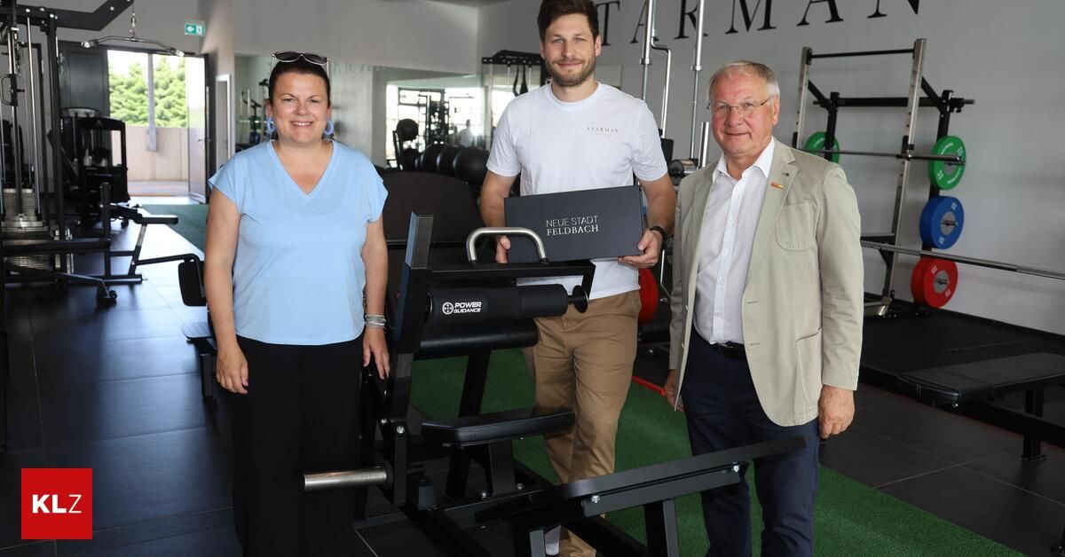 Drei Personen stehen in einem Fitnessstudio. Eine Frau in einem blauen Shirt und ein Mann in einem weißen T-Shirt halten eine Box. Der Mann im beigefarbenen Anzug lächelt.
