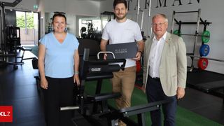 Drei Personen stehen in einem Fitnessstudio. Eine Frau in einem blauen Shirt und ein Mann in einem weißen T-Shirt halten eine Box. Der Mann im beigefarbenen Anzug lächelt.