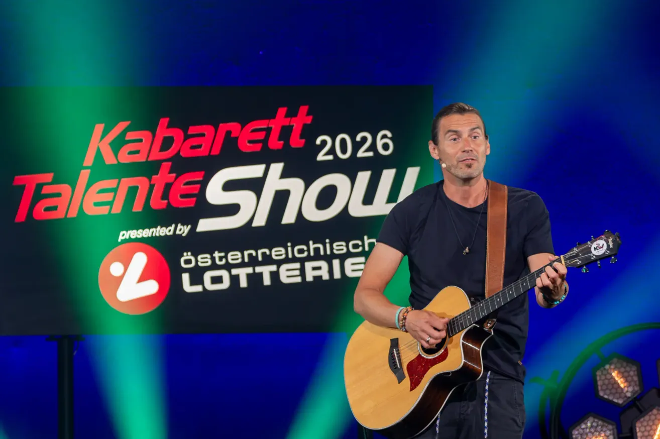 Ein Mann spielt auf der Bühne mit einer Gitarre und trägt ein schwarzes T-Shirt und eine Halskette. Der Hintergrund hinter ihm zeigt ein Logo für Kabarett 2026 präsentiert von Österreichische Lotterie.