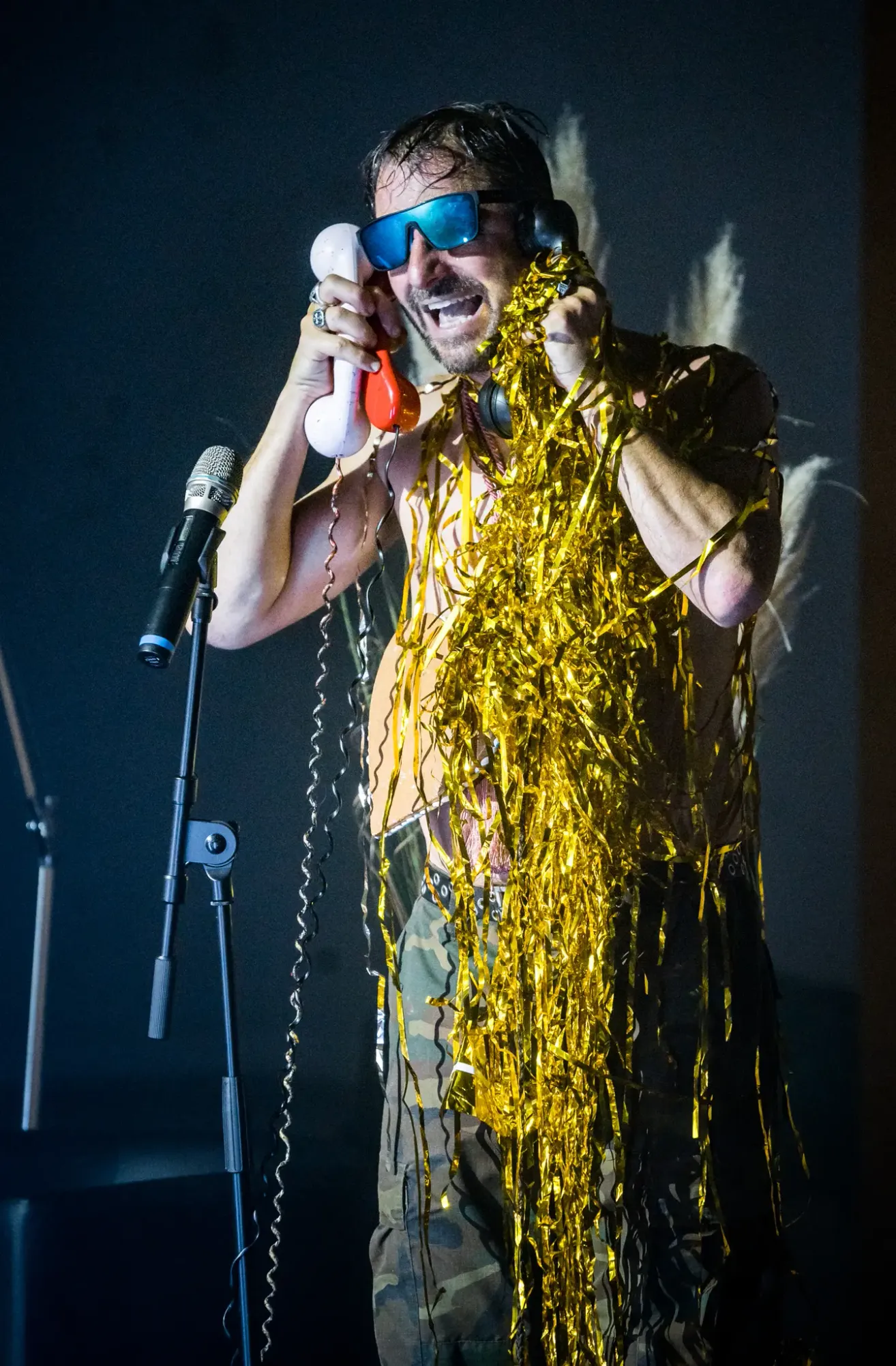 Ein unbekleideter Mann mit goldenen Bändern auf der Bühne hält ein Telefon und spricht in ein Mikrofon.