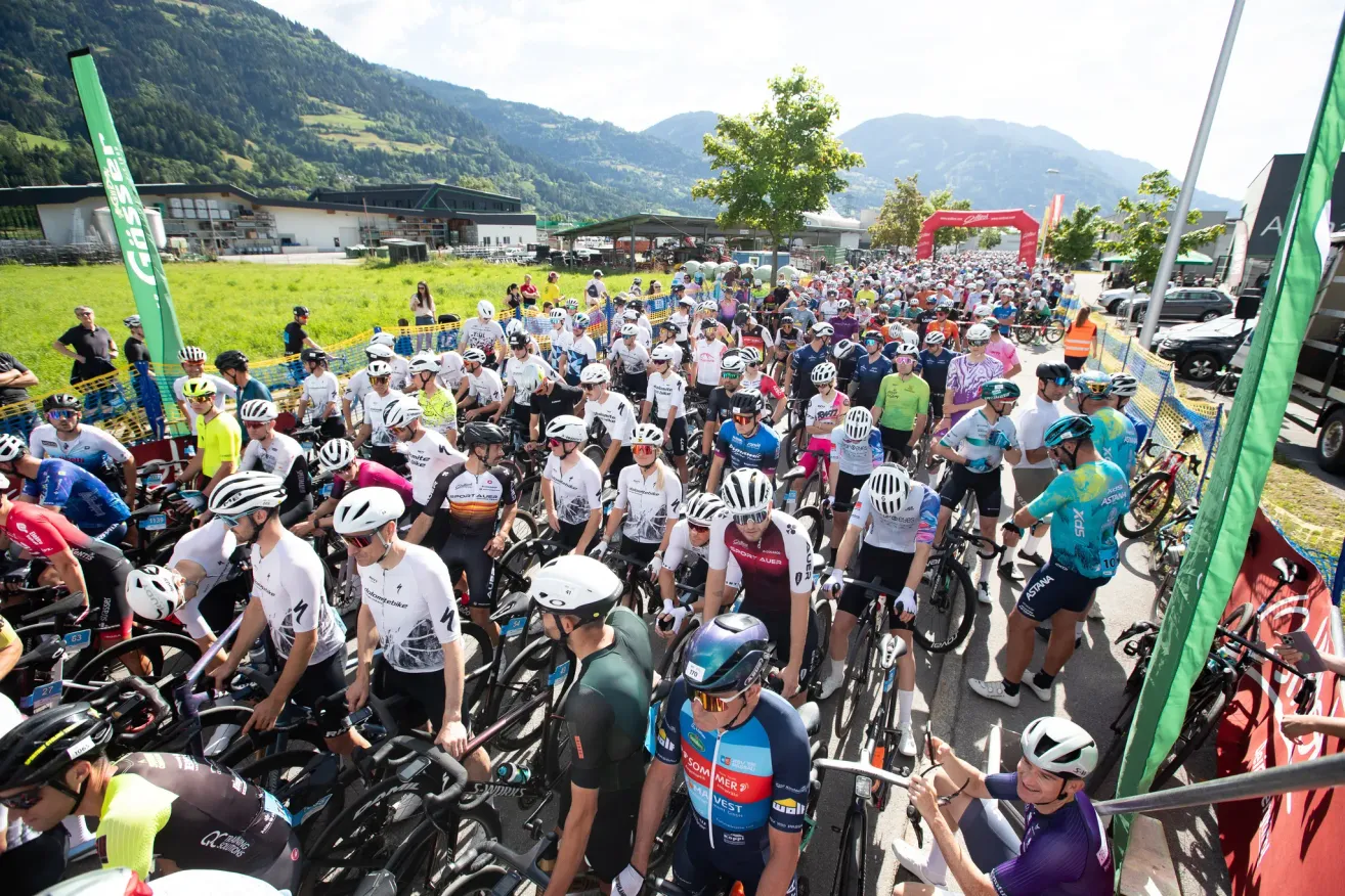 Eine große Gruppe von Radfahrern in Helmen und Radsportausrüstung versammelt sich für ein Rennen in einer malerischen Bergregion. Zuschauer und Gebäude sind im Hintergrund zu sehen.