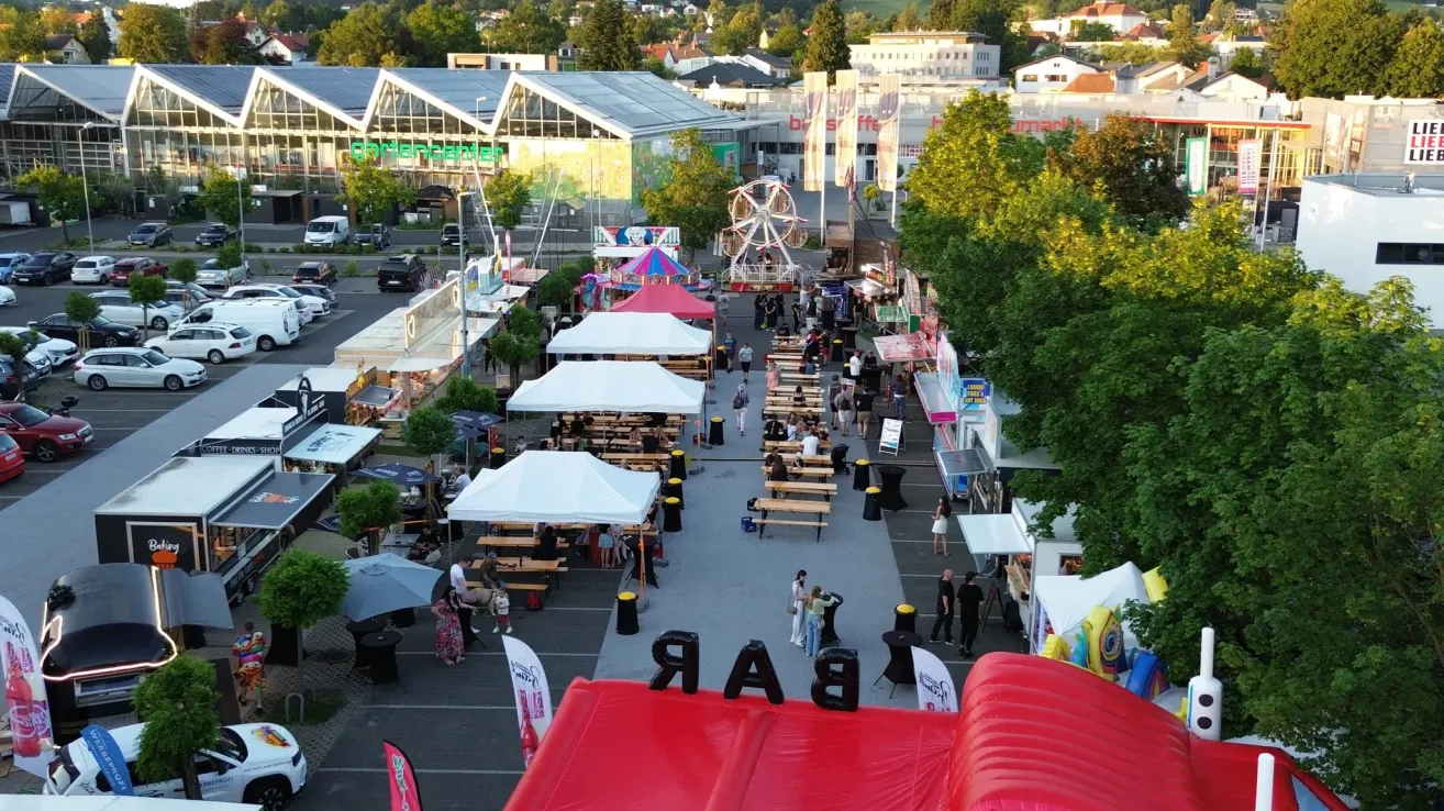 Luftaufnahme eines geschäftigen Freiluftmarktes mit vielen Zelten, Essensständen und einem Riesenrad. Mehrere Menschen gehen herum und sitzen an Tischen, genießen den Jahrmarkt.