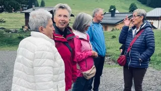 Vier Personen stehen auf einem Kiesweg und plaudern lächelnd. Hinter ihnen steht ein Haus mit einem Schornstein und grüner Wiese.