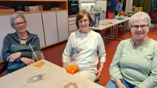 Eine ältere Frau sitzt in einer Küche mit einem Getränk in einem Glas vor ihr. Eine andere Person sitzt zu ihrer Rechten.