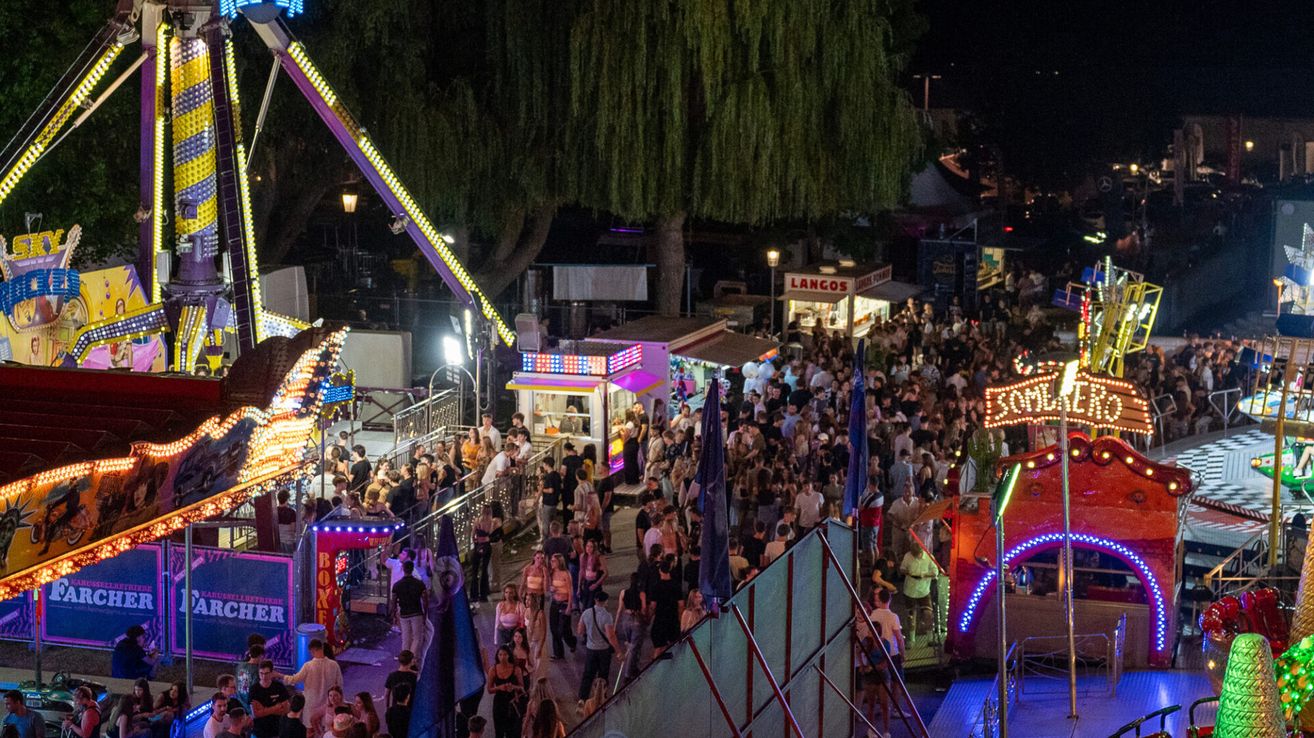 Ein überfülltes Outdoor-Event bei Nacht mit Menschen, bunten Fahrgeschäften und Imbissständen unter Neonlichtern.