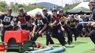 Eine Gruppe von Männern in Uniform rennt auf eine rote Maschine zu, während sie Helme tragen. Sie sind wahrscheinlich Feuerwehrleute. Im Hintergrund steht ein Zelt.