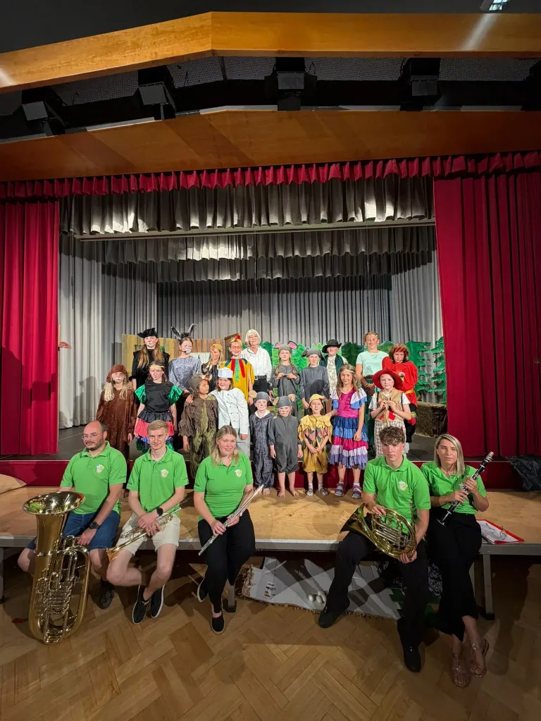 Eine Gruppe von Darstellern in Kostümen, darunter Erwachsene und Kinder, stehen auf einer Bühne mit Musikinstrumenten vor ihnen. Dahinter befindet sich ein roter Vorhang und eine Kulisse.