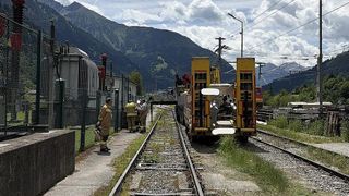 Eine Gruppe von Menschen versammelt sich um eine Eisenbahnschiene, mit einem Zug auf der Seite und einem Berg im Hintergrund.