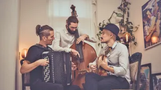 Drei Musiker sitzen auf einem Sofa. Einer spielt Akkordeon, ein anderer Cello und der dritte Gitarre. Sie sehen konzentriert auf ihre Darbietung.