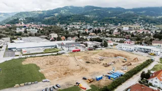 Ein Luftbild von einer Baustelle mit mehreren Fahrzeugen, Maschinen und Gebäuden. In der Nähe befinden sich Berge und eine Stadt mit verschiedenen Häusern.