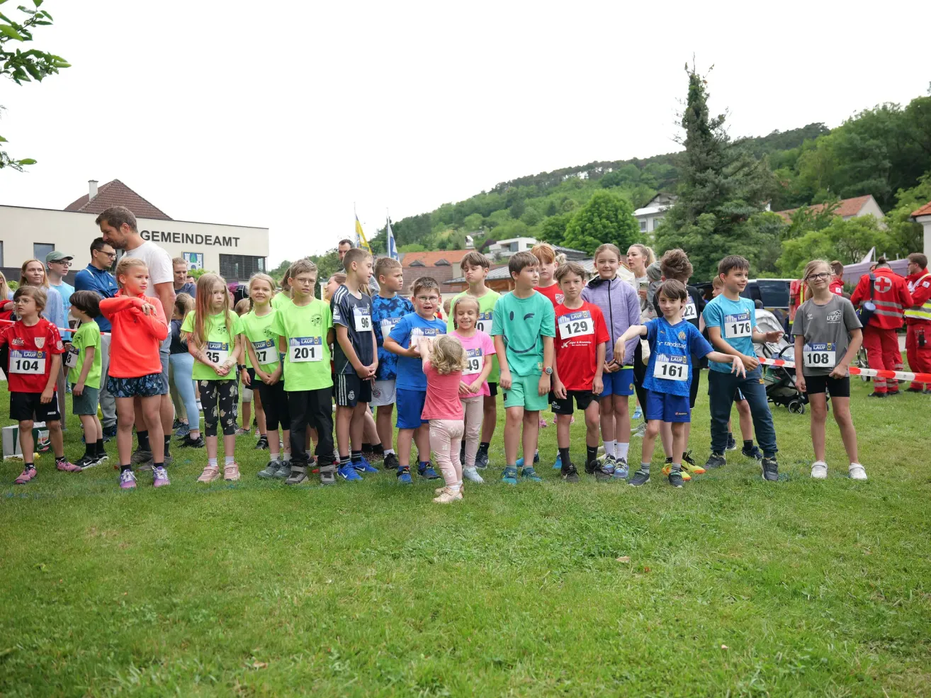 Eine Gruppe von Kindern mit nummerierten Startnummern steht auf einem Rasenfeld vor einem Gebäude. Im Hintergrund sind Bäume und Häuser zu sehen.